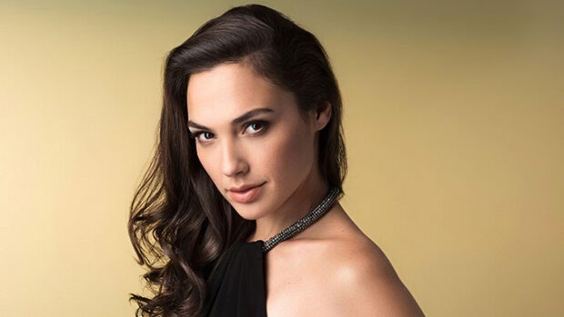 Gal Gadot posing confidently with long dark hair and a black dress against a beige background