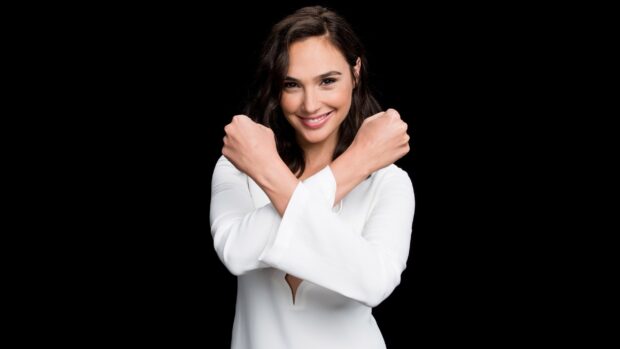 Gal Gadot smiling with crossed arms wearing a white top against black background