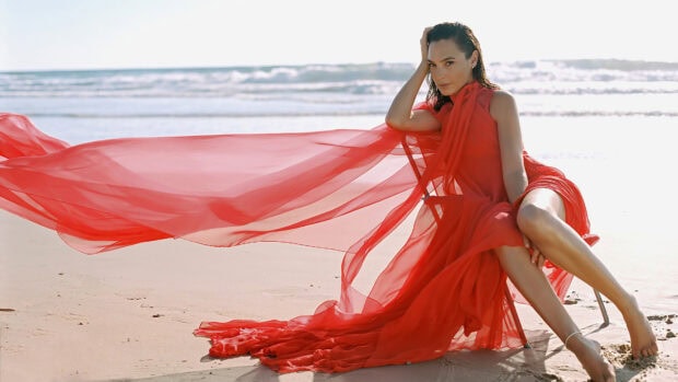 Gal Gadot sitting on the beach wearing a flowing red dress with ocean waves in the background