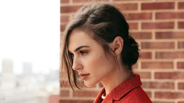 Gal Gadot wearing a red coat standing in front of a brick wall looking thoughtfully to the side