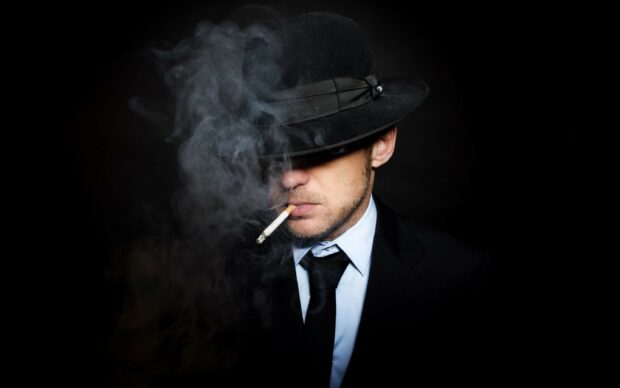 A gentleman in a suit wearing a hat smoking a cigarette with smoke around him