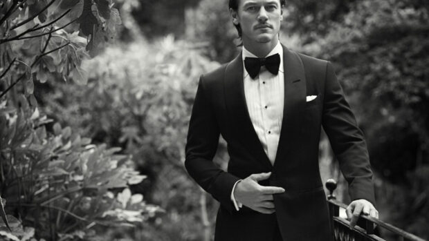 A gentleman wearing a tuxedo stands confidently by a garden railing in black and white photograph