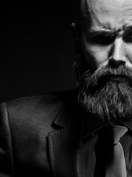 A confident gentleman with a thick beard wearing a suit and tie in a dark setting