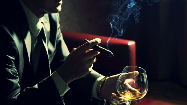 A gentleman holding a cigar and a glass of whiskey in a suit and tie