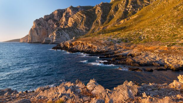 Rocky coastal cliffs under golden sunlight at the edge of the sea with clear blue water