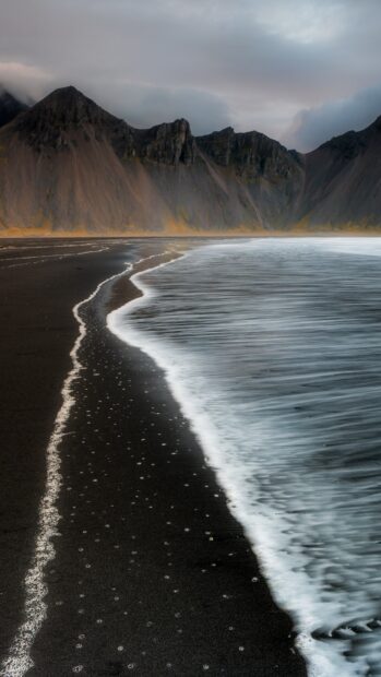 A volcanic black sand beach with waves washing ashore near rocky coastline geography