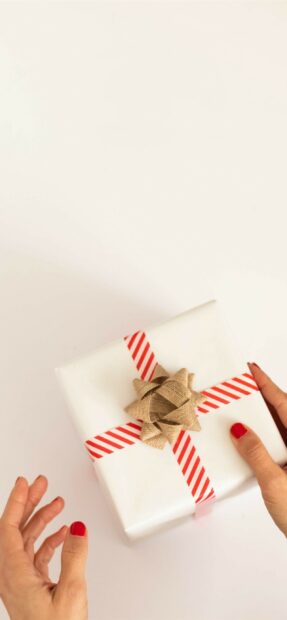 Person holding a white gift with striped tape and a rustic bow on a plain surface