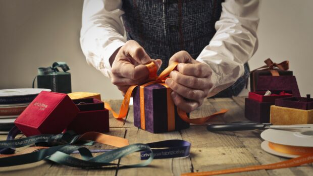 A person tying a ribbon around a gift box on a wooden table surrounded by other gift boxes and ribbons