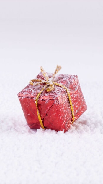 Red gift wrapped in shiny paper with golden ribbon covered in snow on white surface
