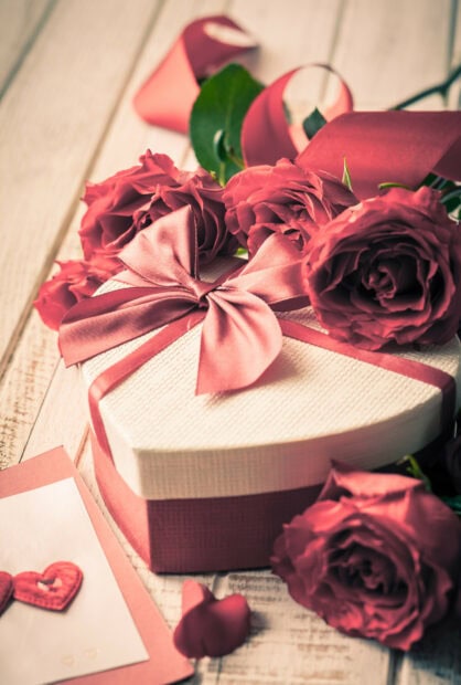 A heart shaped gift box with a pink ribbon and roses on a wooden table