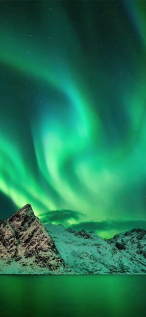 Green aurora lights swirl over snow covered mountains and calm water at night