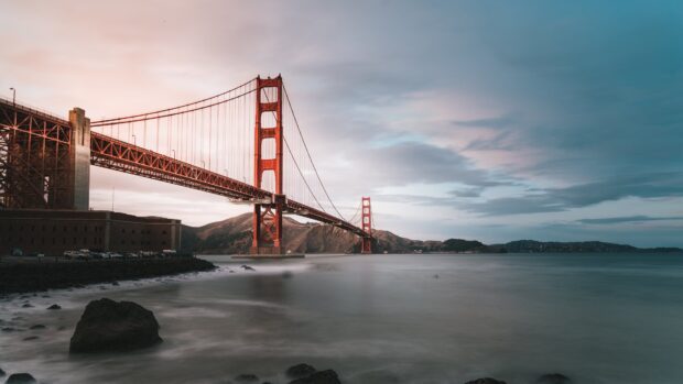 Golden Gate Bridge is seen from the rocky shore with hills in the background at dusk