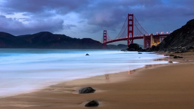 A view of Golden Gate Bridge at dusk with waves gently hitting the sandy beach and rocky shoreline