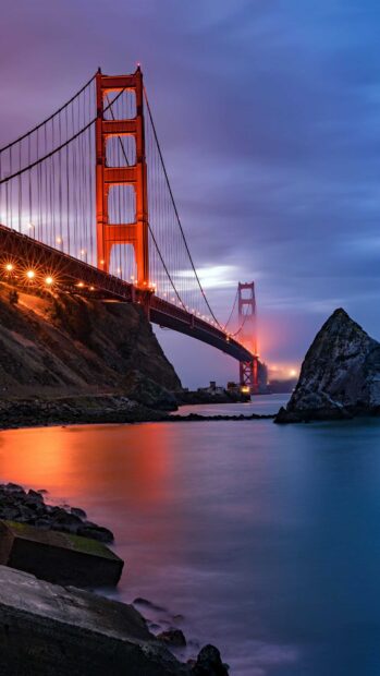 Golden Gate Bridge at dusk with vivid lights and fog over the water viewed from the rocky shore