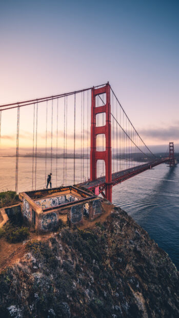 Person walking near Golden Gate Bridge on cliffside at sunset with city in background