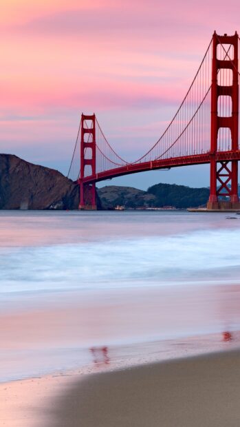 The Golden Gate Bridge at sunset with calm waves on the beach and hills in the background