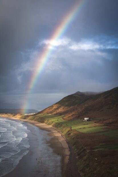 A serene beach and hillside landscape with a rainbow over the Gower Peninsula