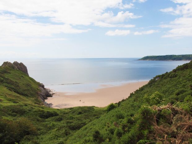 A scenic view of the peninsula with lush green hills meeting a sandy beach and calm sea in Gower Peninsula