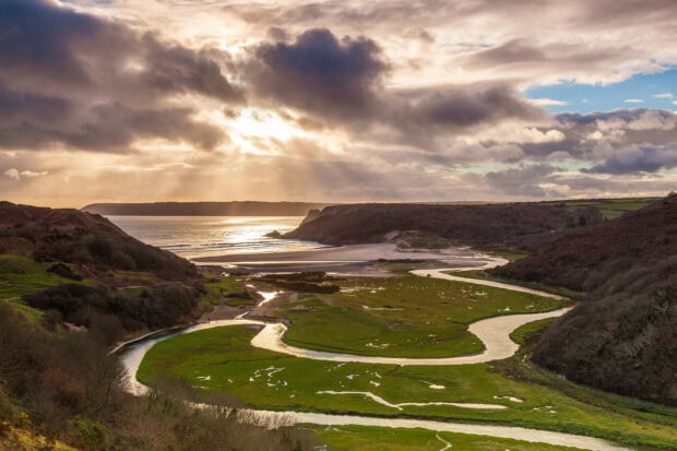 A winding river flowing through green hills on Gower Peninsula under a dramatic cloudy sky