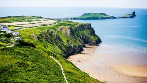 Green cliffs and sandy beach along Gower Peninsula coastline in bright sunlight