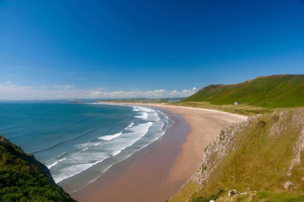A scenic coastline of Gower Peninsula with rolling hills and sandy shore under a clear blue sky