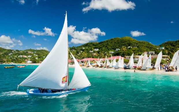 A traditional Grenada sailboat gliding through turquoise waters near a lively beach with hills in the background