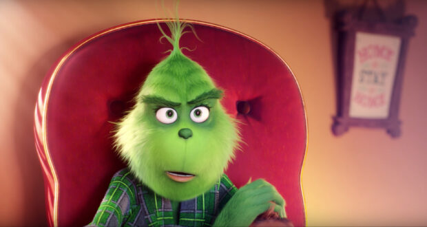 The Grinch with green fur sitting on a red chair wearing a plaid shirt looking surprised