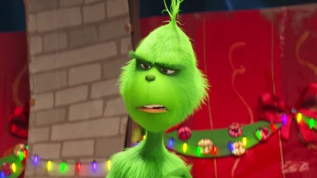 The Grinch character with green fur and expressive face standing in front of colorful Christmas decorations