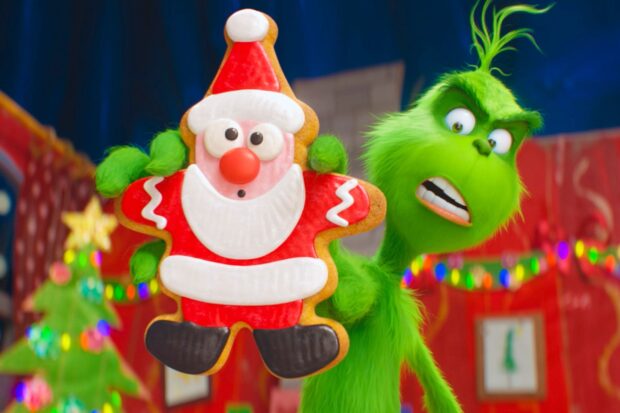The Grinch holding a large Santa shaped cookie with colorful Christmas lights in the background