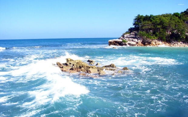 Waves crashing against rocky shore in Haiti sea with clear blue sky and green cliffs