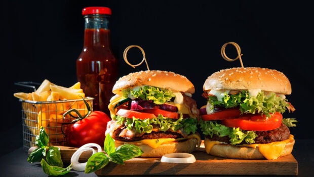 Two beef hamburgers with lettuce tomato cheese and bacon served with fries and ketchup sauce on wooden board