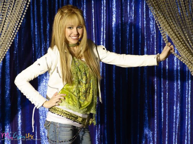 A smiling Hannah Montana posing in a white jacket and green top against a blue curtain background