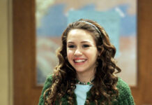 A young woman with curly hair wearing a green knitted cardigan and floral skirt smiling indoors