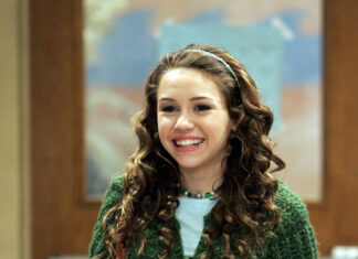 A young woman with curly hair wearing a green knitted cardigan and floral skirt smiling indoors