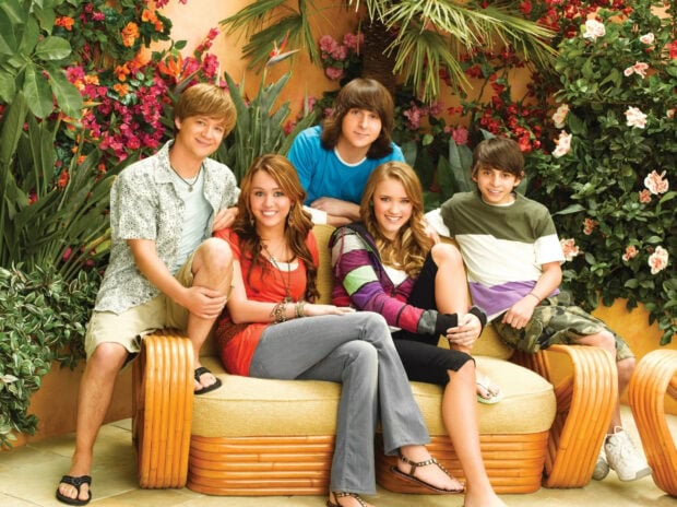 The Hannah Montana cast sitting together on a sofa surrounded by vibrant flowers and greenery