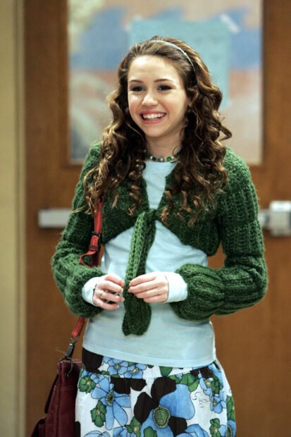 Teenage girl with curly hair wearing a green sweater and floral skirt smiling brightly