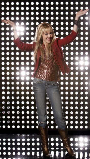 Hannah Montana wearing a red jacket and jeans posing with arms raised against bright lights background