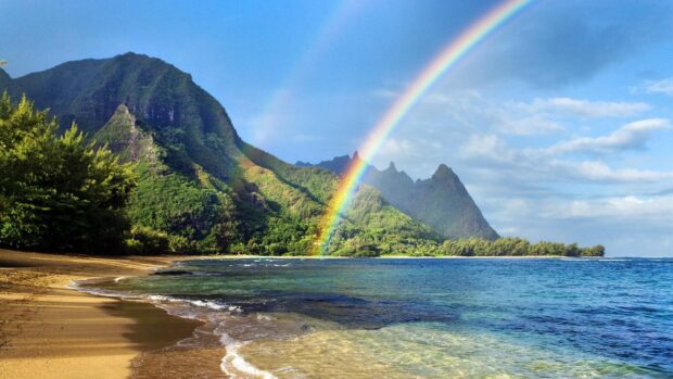 A scenic view of Hawaiian Islands with a rainbow over lush green mountains and a sandy beach