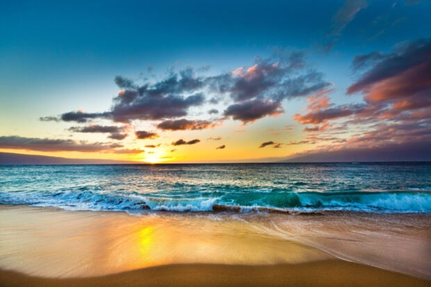 Hawaiian ocean waves gently crashing on the sandy beach at sunset with colorful clouds in the sky