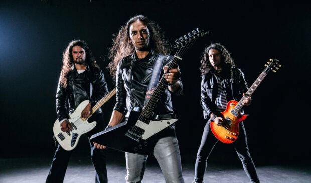 Three men in leather jackets playing guitars in a heavy metal band setting