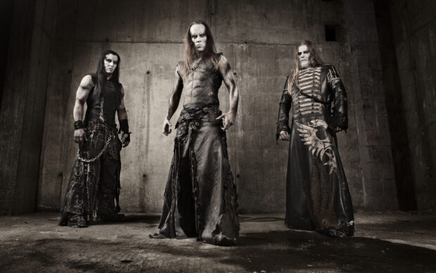 Three heavy metal musicians wearing dark gothic outfits in an industrial setting
