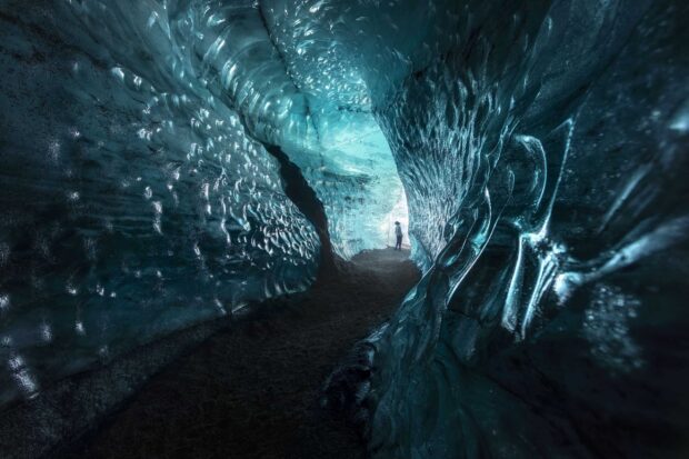 A person exploring the vast ice cave interior with textured frozen walls and glowing light ahead