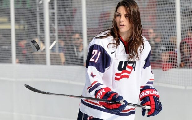 Female ice hockey player in USA jersey holding hockey stick with puck in midair