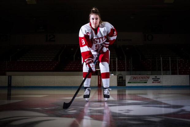 Female ice hockey player wearing Boston jersey poses on ice hockey rink with stick