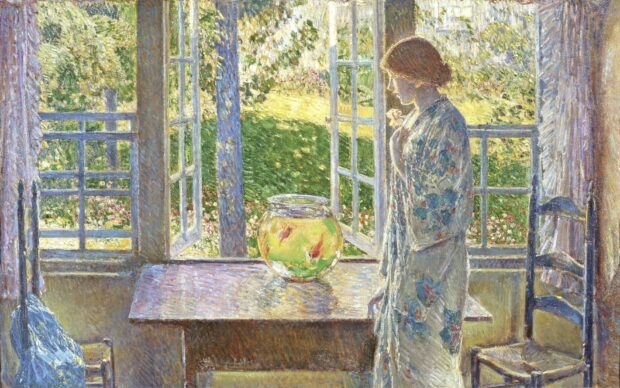 Woman in a floral robe standing by a table with a goldfish bowl near open windows with garden views