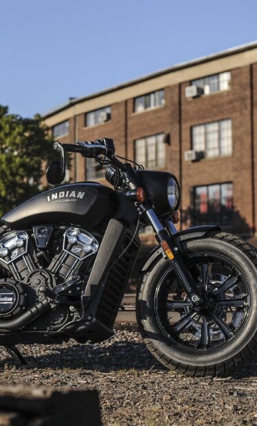 Black Indian Scout bike parked on rough ground with a brick building in the background