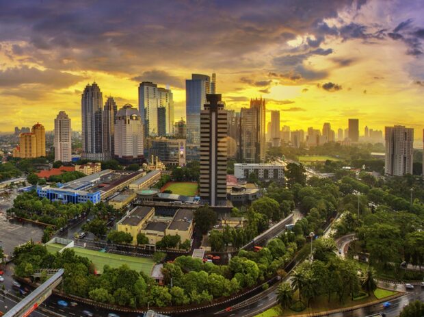 A cityscape of Indonesia featuring tall buildings and lush greenery at sunset with dramatic clouds
