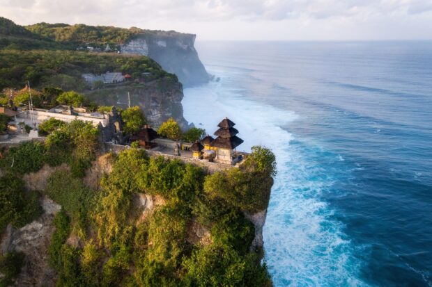 Ancient Indonesian temple on cliff surrounded by lush greenery overlooking the ocean waves