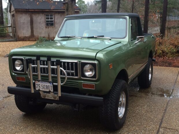 Green International Harvester Scout truck parked on driveway with wooden cabin in background