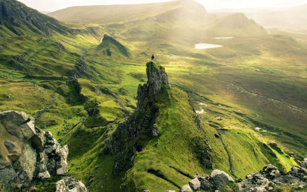 A lone hiker stands on a rocky ridge overlooking the vast Isle of Skye landscape in vibrant green tones